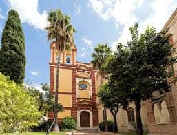Vista exterior de la Iglesia de San Agustín con fachada amarilla y detalles rojos, rodeada de palmeras y árboles