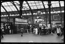 Interior histórico de la estación con viajeros y reloj colgante en blanco y negro