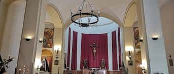 Interior del altar principal de la parroquia, con cortinas rojas, crucifijo central y arcos blancos que dan luz al espacio sagrado