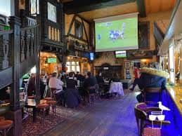 Interior del pub con barra, mesas y pantalla mostrando un partido de fútbol