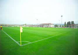 Campo de césped de Tajonar con banderines de esquina y edificios deportivos al fondo