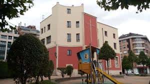 Fachada lateral de la parroquia con tonos beige y rojos, rodeada de árboles y un parque infantil