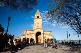 Plaza frente a la iglesia con personas caminando y árboles bajo cielo azul