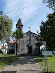 Vista exterior de la Casa do Pobo de Cerponzóns, un edificio de piedra con torre y cruz en la fachada, rodeado de árboles y césped