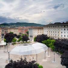 Perspectiva elevada de la plaza mostrando el paraguas blanco y el entorno urbano con edificios y montañas al fondo