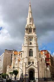 Vista exterior de la iglesia con su característica torre y reloj, bajo nubes dramáticas