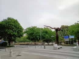 Plaza abierta con escultura moderna y bancos bajo árboles