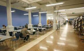 Interior amplio de la biblioteca con mesas de estudio, columnas blancas y estanterías de libros