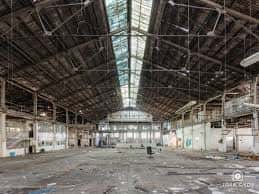 Interior del nave principal de la fábrica con estructura metálica y techo de cristal, mostrando el espacio amplio y abandonado
