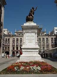 Monumento a Pedro Velarde en el centro de la plaza con jardines floridos y edificios históricos al fondo