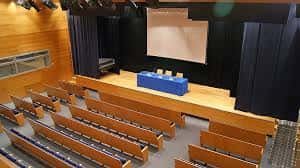 Interior del auditorio con escenario, pantalla y butacas de madera