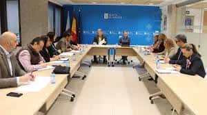 Sala de juntas con mesa rectangular y participantes en reunión institucional con banderas de Galicia y España