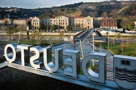 Letrero 'DEUSTO' en primer plano con el campus universitario y el río Nervión al fondo