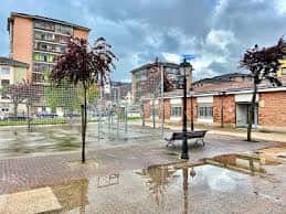 Vista exterior del centro social con plaza adoquinada, árboles y bancos bajo cielo nublado