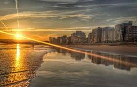 Atardecer sobre la playa de San Lorenzo con reflejos en la arena mojada y skyline urbano