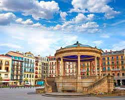 Kiosco de la Plaza del Castillo bajo un cielo azul con nubes, rodeado de edificios coloridos