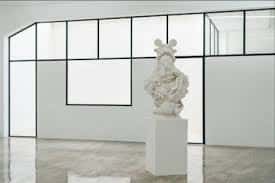 Detalle de una escultura blanca sobre pedestal en la Galería Fermay con fondo de ventanales