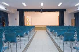 Sala de actos con butacas azules y escenario con cortinas oscuras