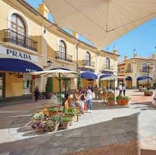Plaza interior del outlet con tiendas como Prada, sombrillas y zonas de descanso al aire libre