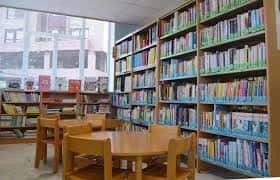 Interior de la biblioteca con estanterías llenas de libros y mesas de lectura junto a ventana