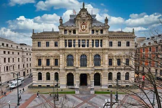 Vista frontal del Palacio Foral de Bilbao con cielo azul y nubes, mostrando su fachada neoclásica y plaza delantera