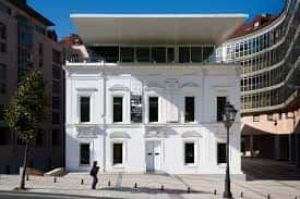 Entrada principal del edificio del Colegio Oficial de Arquitectos de Asturias, con detalle de la fachada blanca y elementos urbanos circundantes