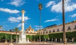 Plaza de la Constitución con el monumento a la Constitución y palmeras bajo un cielo azul
