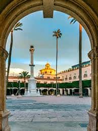 Vista de la Plaza de la Constitución enmarcada por un arco, con el monumento y palmeras al fondo