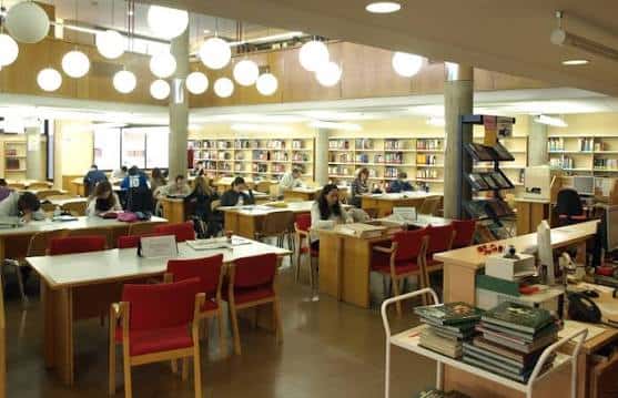 Interior de la biblioteca con mesas de estudio, sillas rojas y estanterías llenas de libros