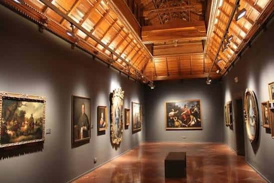 Interior de una sala de exposiciones con cuadros históricos y techo de vigas de madera