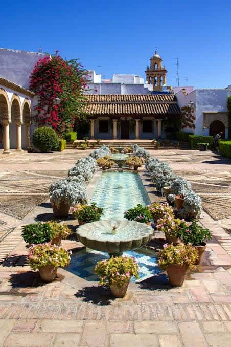 Vista amplia del patio con fuente, estanque y fachada del palacio bajo cielo azul