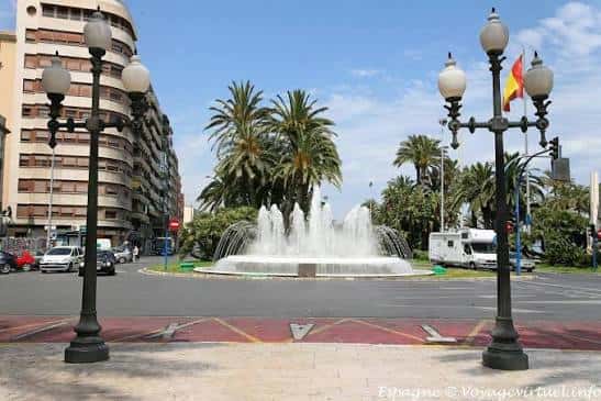Vista frontal de la plaza con la fuente iluminada y palmeras