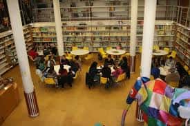 Interior de la biblioteca con mesas redondas, estanterías y personas leyendo en ambiente tranquilo