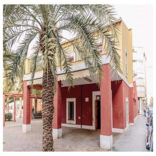 Entrada del centro social con columnas rojas y palmera en primer plano