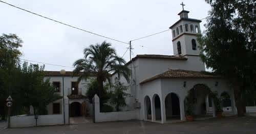 Vista general de la Ermita de Sanz rodeada de palmeras y árboles en un día nublado