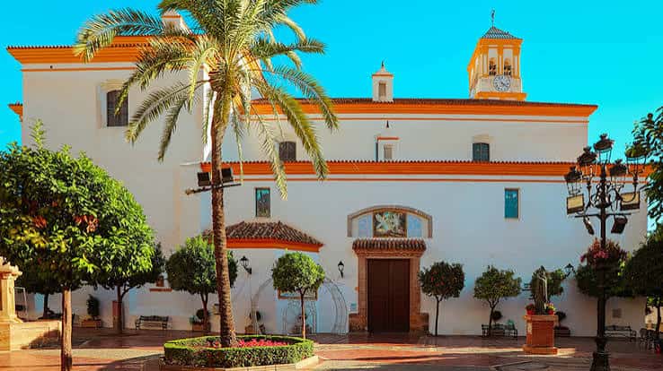 Vista exterior de la iglesia con palmeras y cielo azul, mostrando su arquitectura blanca y campanario
