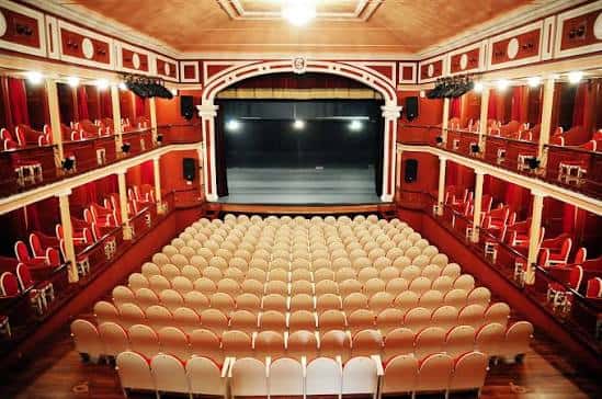 Vista panorámica del interior del Teatro Salón Cervantes con butacas beige y balcones rojos