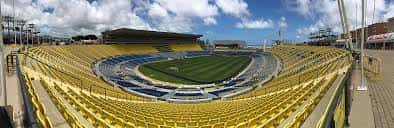 Vista panorámica del interior del estadio con gradas amarillas y césped verde bajo cielo azul