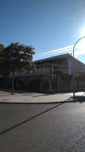 Fachada lateral del complejo deportivo bajo luz solar directa, con árboles y farola en primer plano