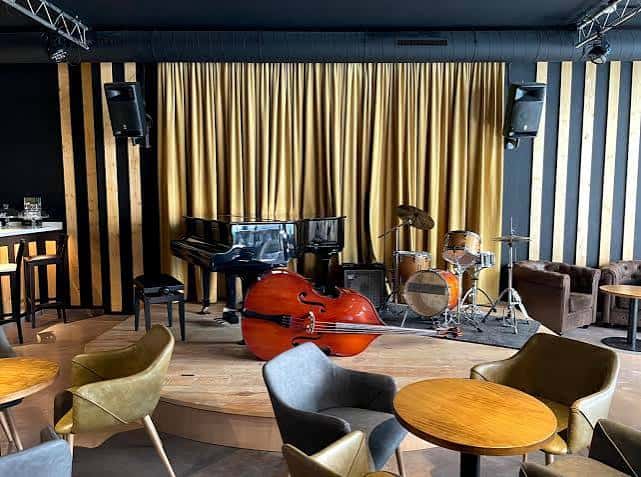 Escenario principal del Cafè de l'Aula con piano de cola, contrabajo y batería, rodeado de mesas y butacas modernas