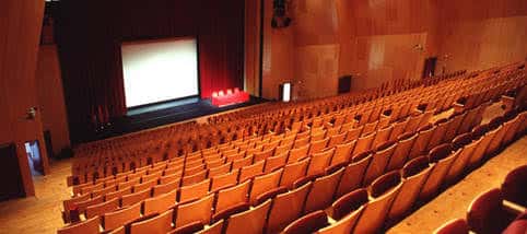 Interior del auditorio mostrando las filas de butacas naranjas y el escenario con pantalla de proyección