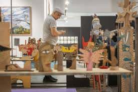 Detalle de una exposición con obras de arte en madera y un artista trabajando en el fondo
