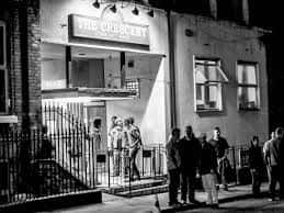 Vista nocturna en blanco y negro de la fachada del venue con personas en la entrada