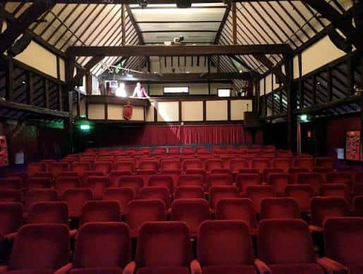 Interior del auditorio con butacas rojas y escenario con cortina roja, bajo una estructura de vigas de madera