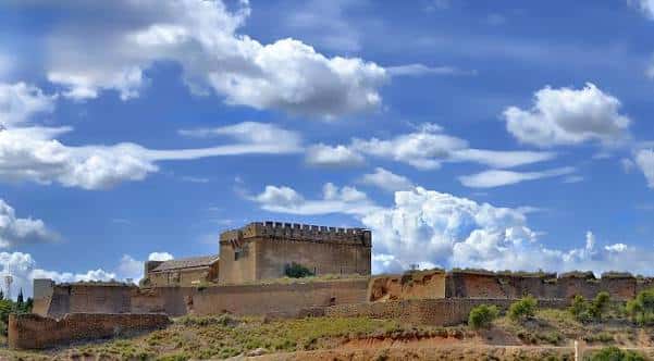 Panorámica del castillo sobre colina con murallas y cielo dramático con nubes blancas