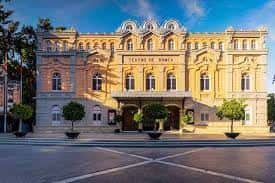 Vista frontal del Teatro Romea al atardecer, con iluminación cálida y árboles decorativos