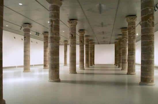 Interior del espacio expositivo principal con columnas de piedra y suelo blanco
