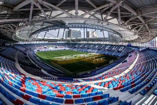 Interior del estadio con estructura metálica del techo y asientos en azul y rojo, mostrando la capacidad y diseño arquitectónico