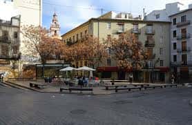 Vista diurna de la Plaza del Tossal con árboles y bancos, mostrando su ambiente urbano y tranquilo