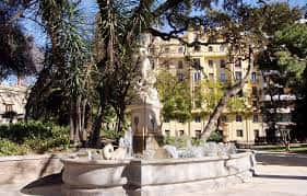 Fuente ornamental con escultura central rodeada de árboles y edificios históricos al fondo